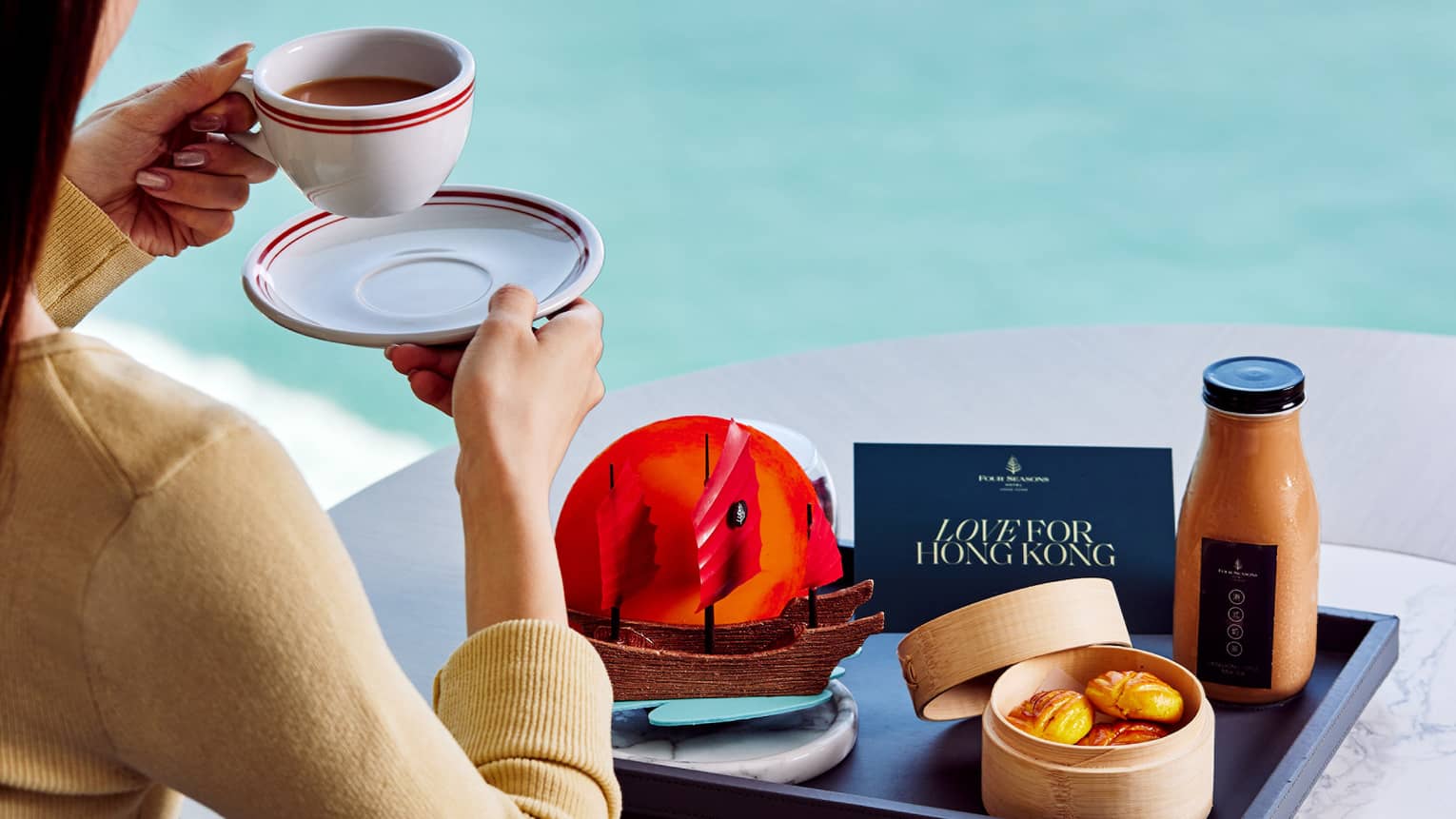 A guest sitting at a table with a bottle of juice, pastries, an edible ship and a card on a tray, while drinking coffee.