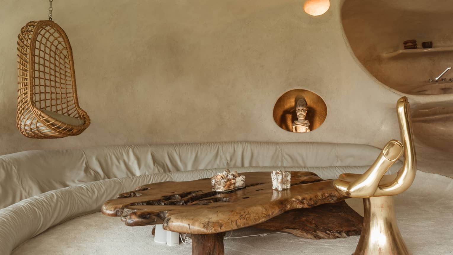 All off-white room with curved walls and unusual furnishings including a couch lining one wall and a gold hand-shaped chair.
