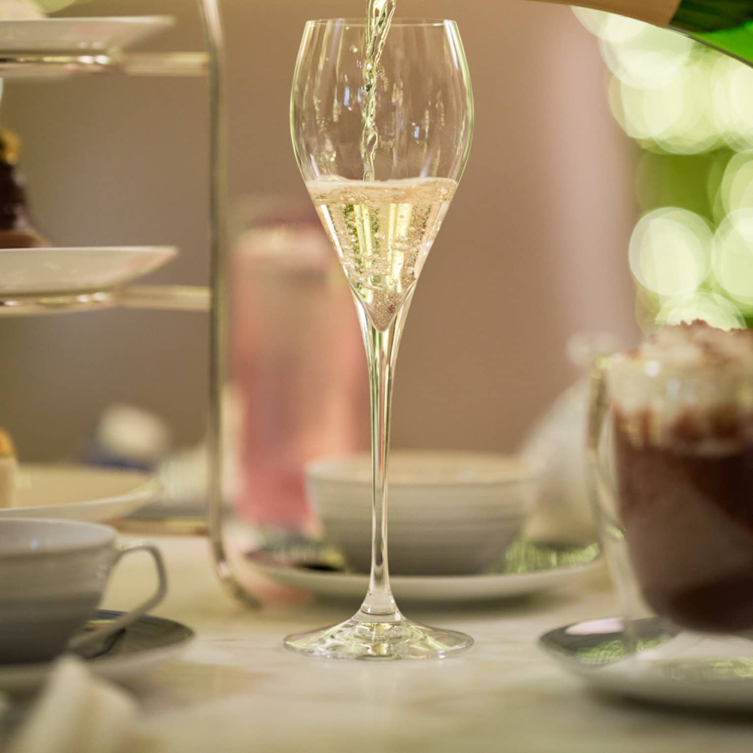 Champagne is poured from a bottle of Perrier Jouet into a glass next to a tiered tray of treats and a frothy cup of espresso.