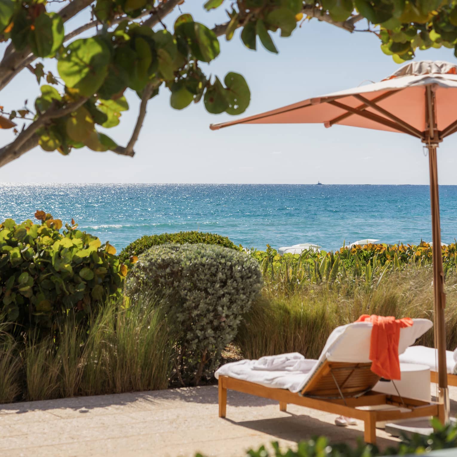 Two lounge chairs set under a pink umbrella sit under a tree with with ocean in the near distance
