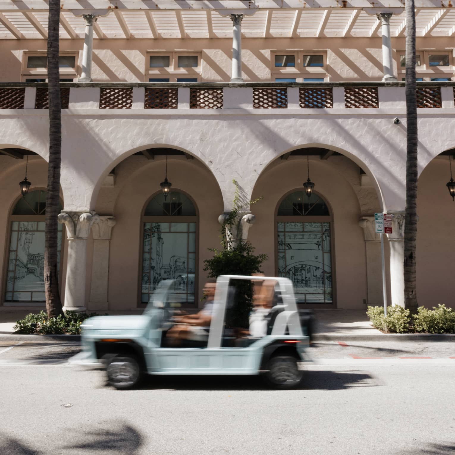 White open-top vehicle drives by a Spanish-style stucco building with palm trees in front