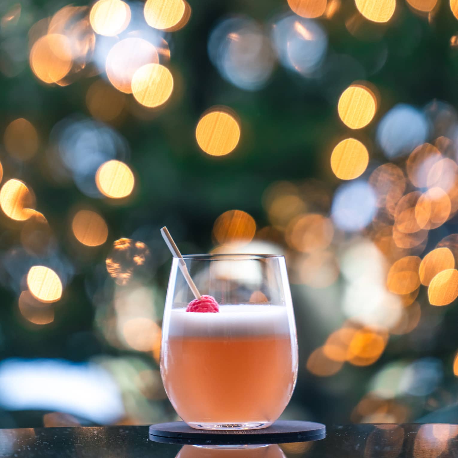An orange-pink coloured cocktail sitting on a table with Christmas lights behind it.
