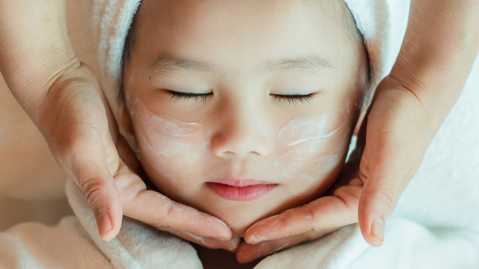 A close-up of a relaxed child wrapped in a towel and robe and enjoying a masseuse massaging a cream into their face.