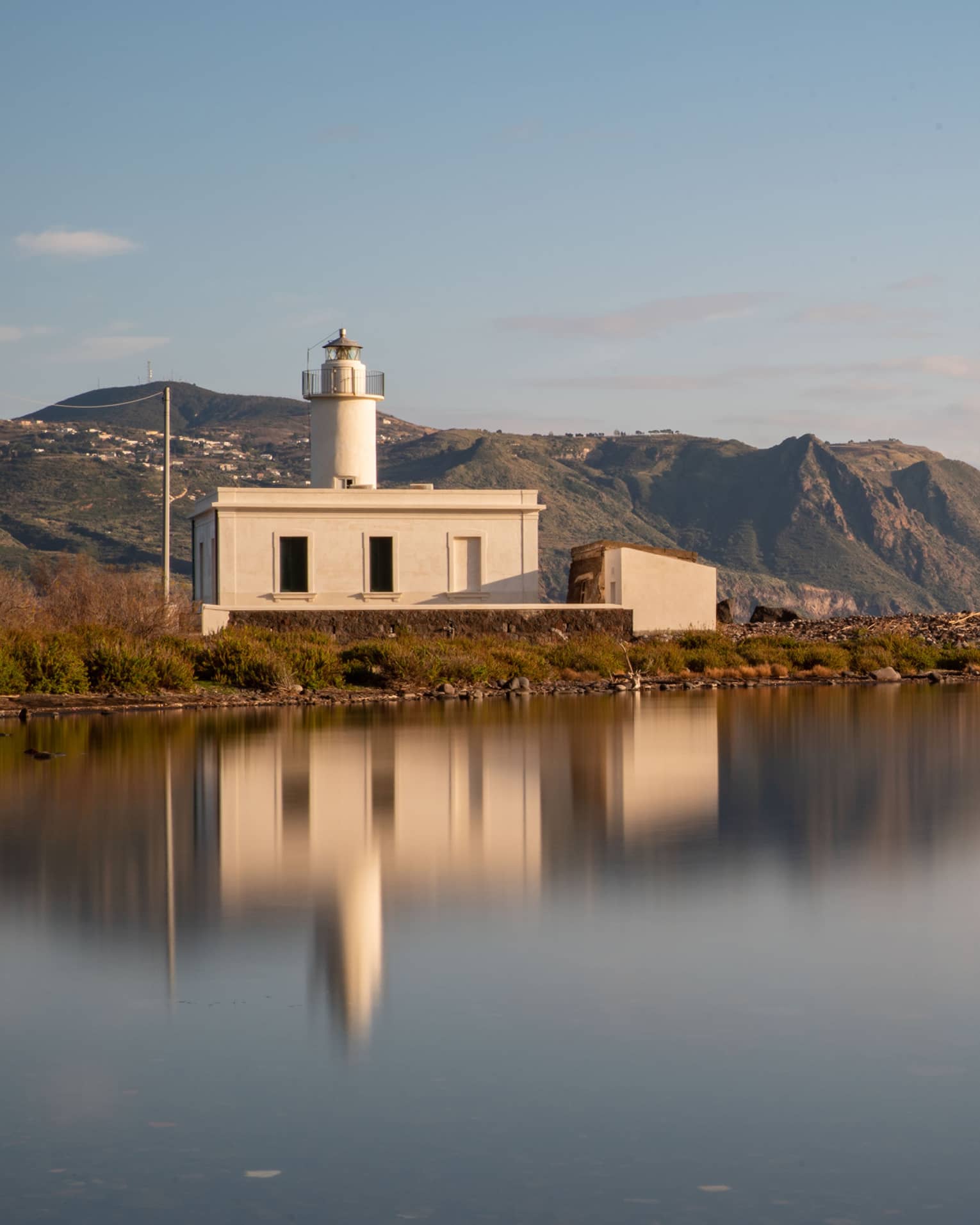 Short all-white lighthouse, reflecting on smooth water, mountains and blue sky in backdrop