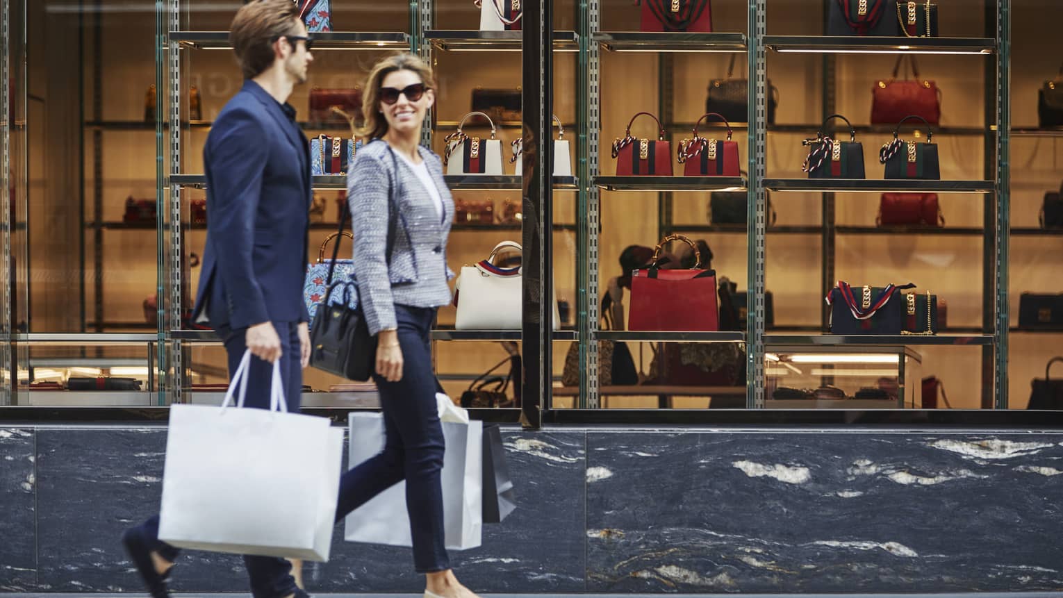 Man and woman in suit jackets, sunglasses hold shopping bags, walk by large store window with designer purses on display 