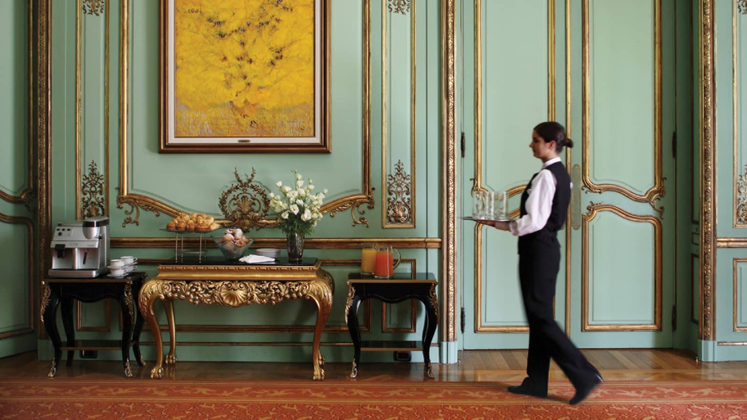 Server in black vest, pants carries tray down historic hallway with gold trim, table