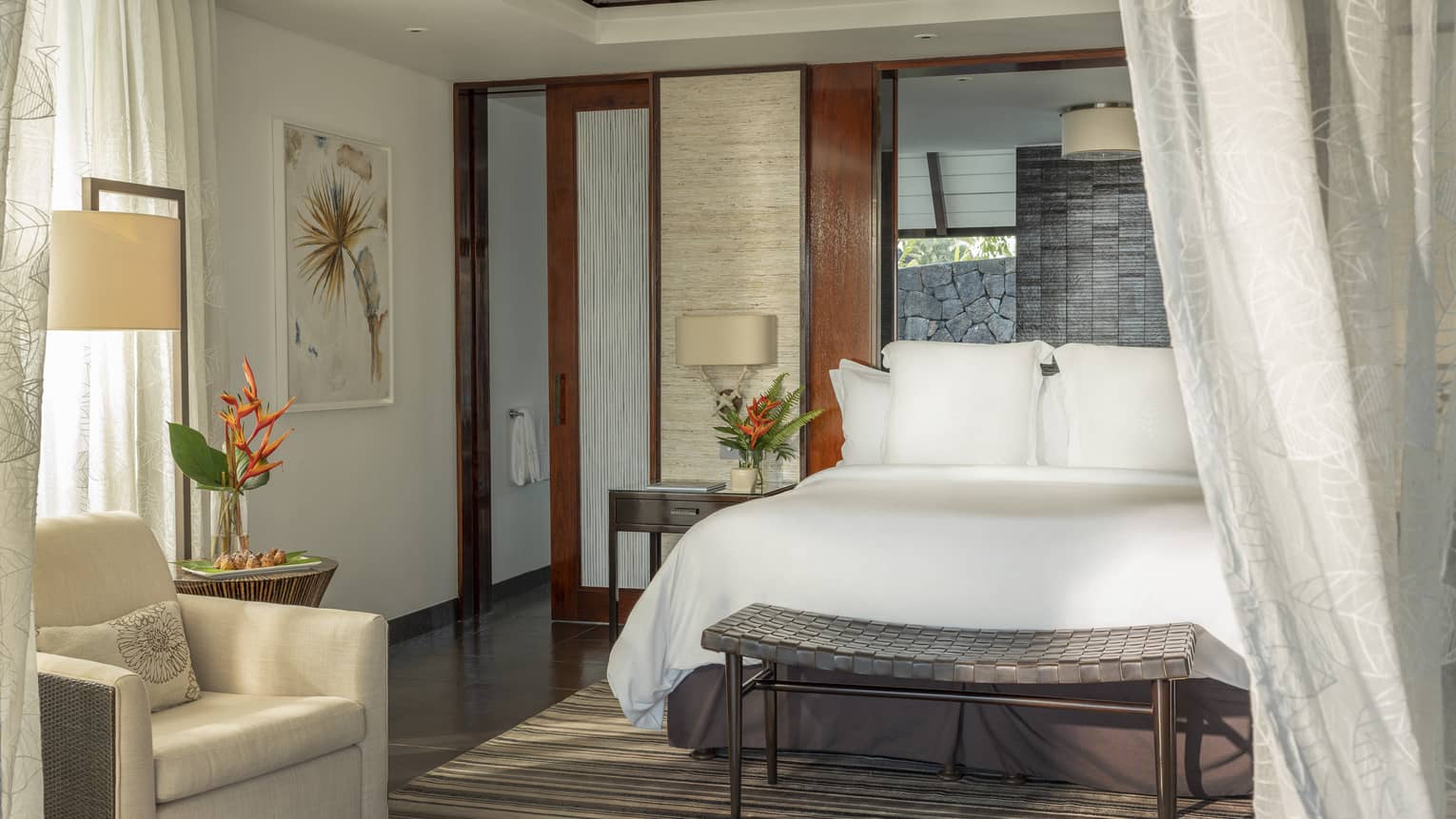 Bedroom with white king bed, whicker bench, long curtains, arm chair