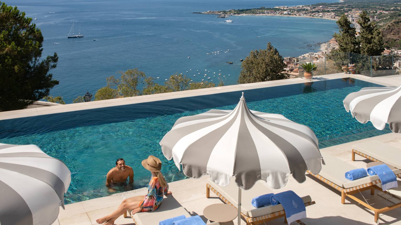 A man at the edge of the infinity pool speaking to a woman sitting on a deck chair