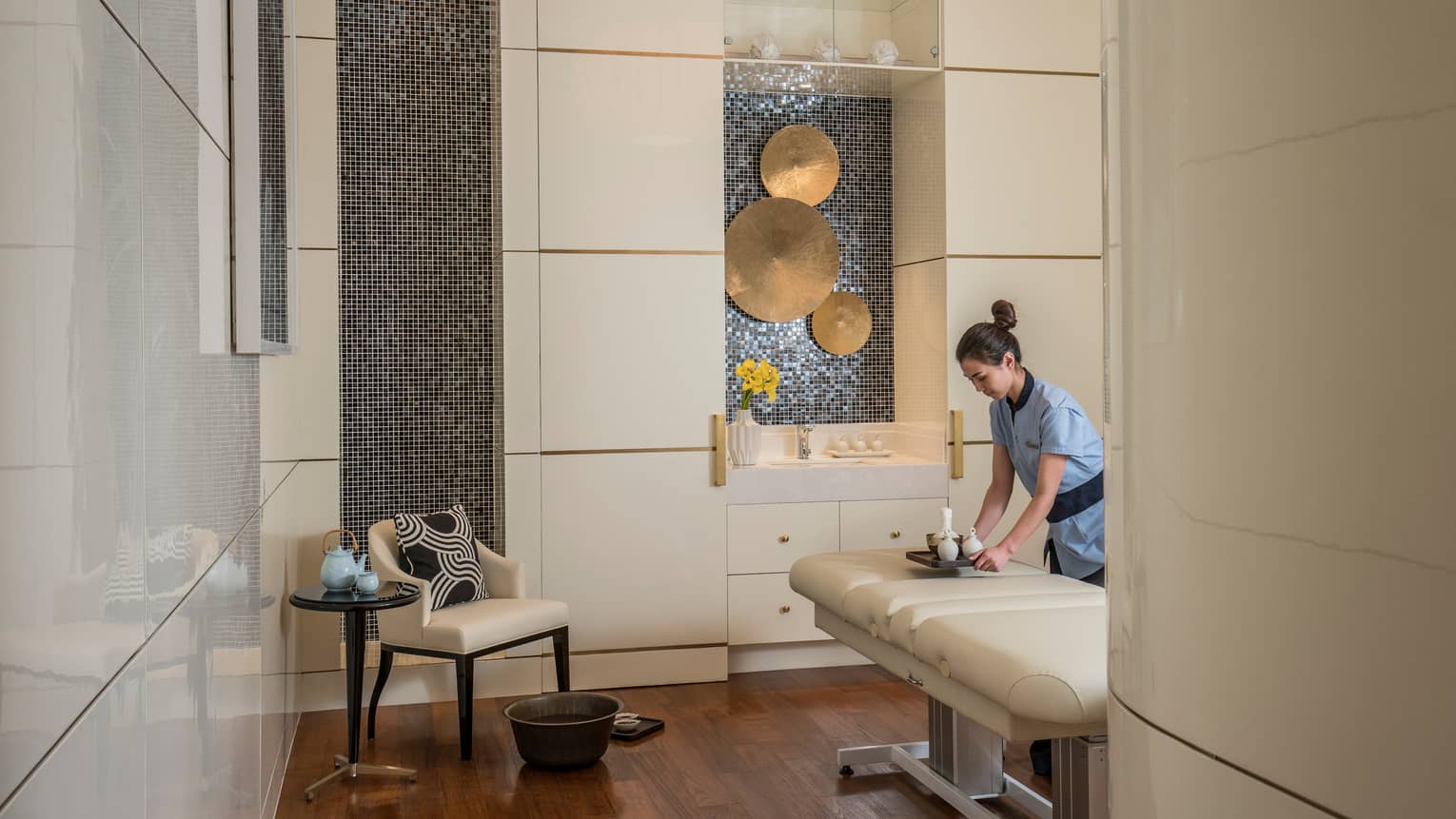 Spa staff sets tray with vases on padded massage table by chair, brass discs on wall