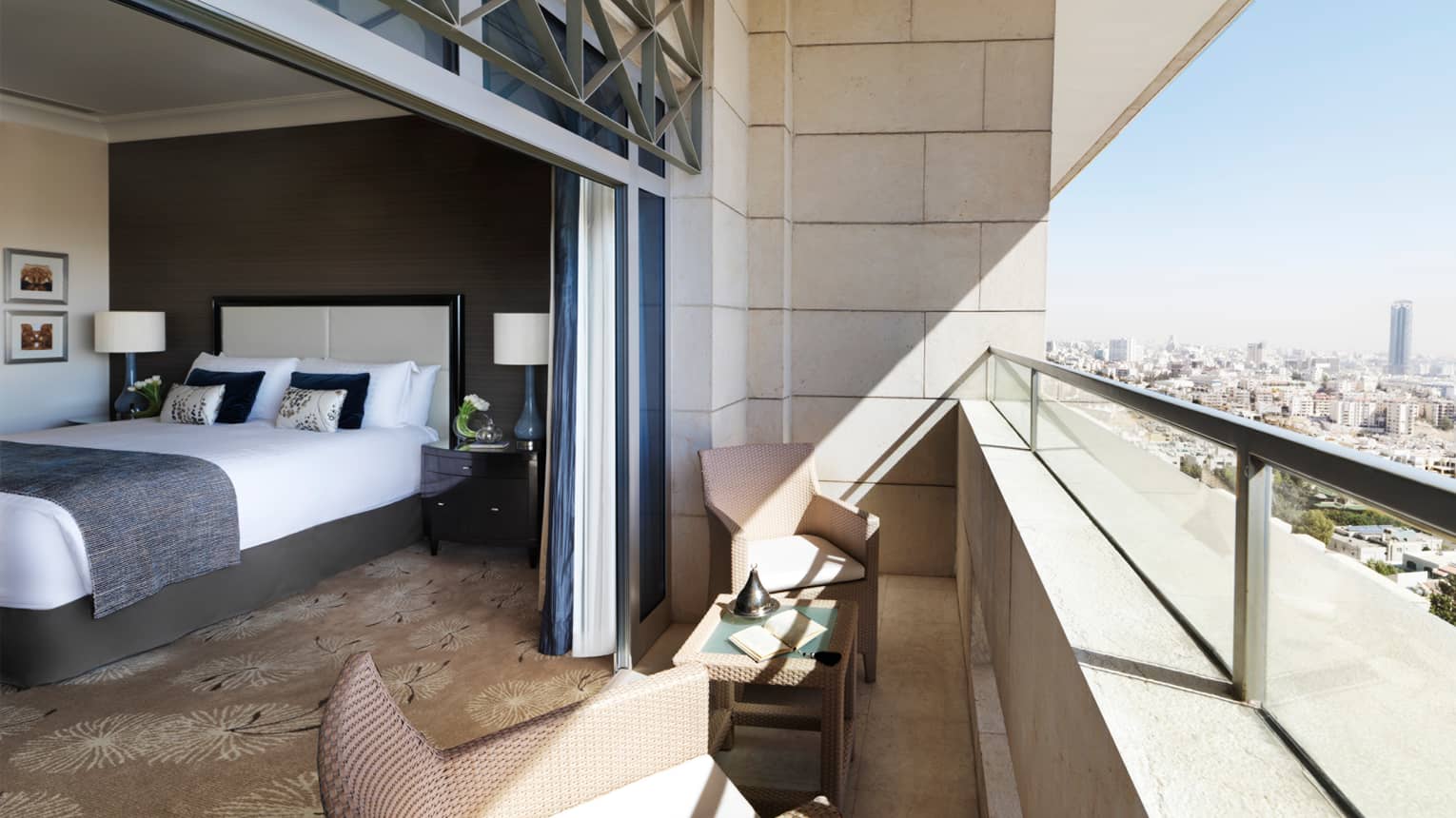 Hotel bedroom with white-and-grey walls, grey blanket on bed, open wall to balcony and city view