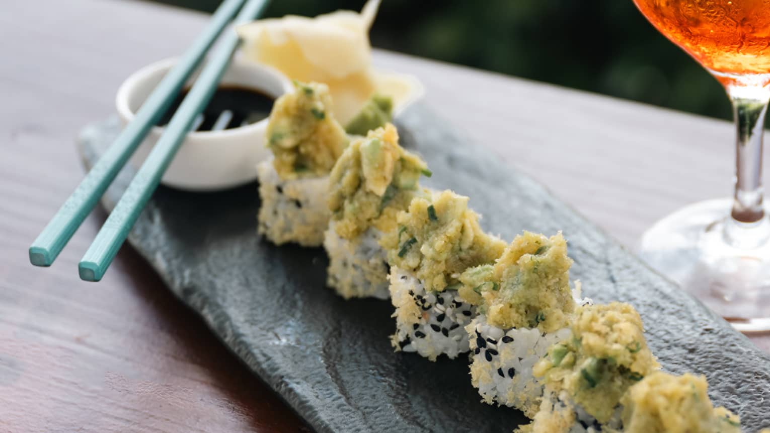 Sushi rolls on tray with chopsticks and wine glass.