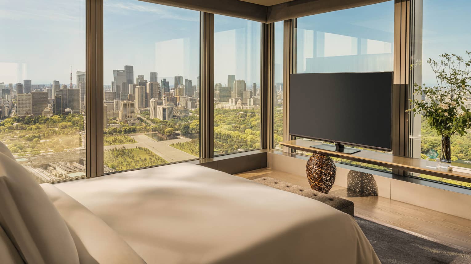 Luxury hotel room featuring king bed, TV and two walls of windows looking out to city and gardens