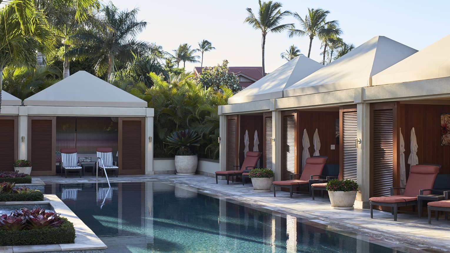 Cabanas line outdoor swimming pool deck under palm trees and sunny sky