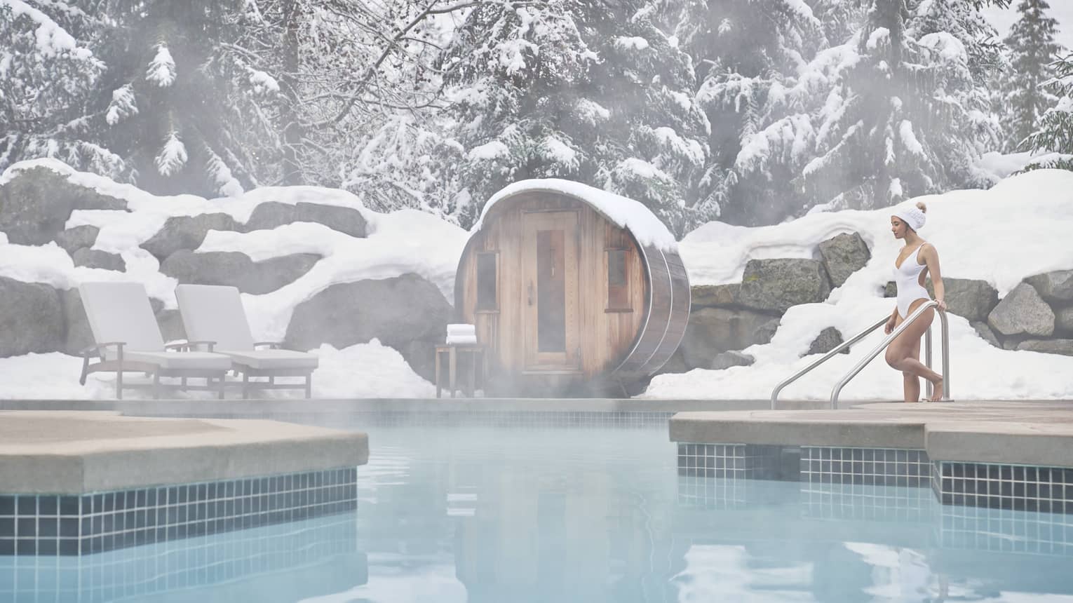 A woman in a white swimsuit enters the outdoor heated pool as steam rises around her. She is surrounded by snow-capped trees.