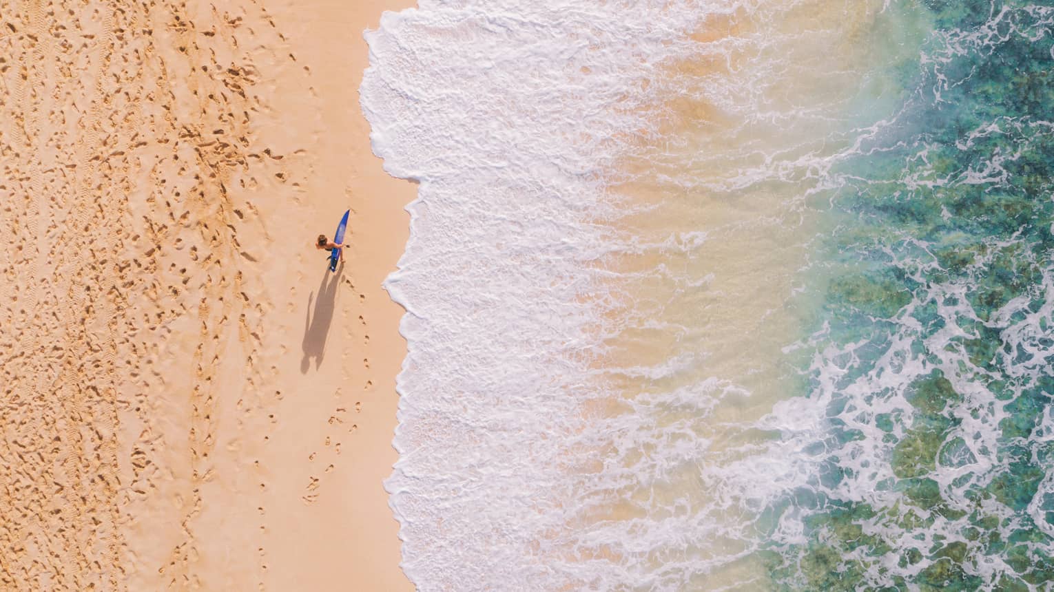 Aerial view of a surfer on a gold-hued beach carrying their board along the edge of waves foaming out of turquoise water.