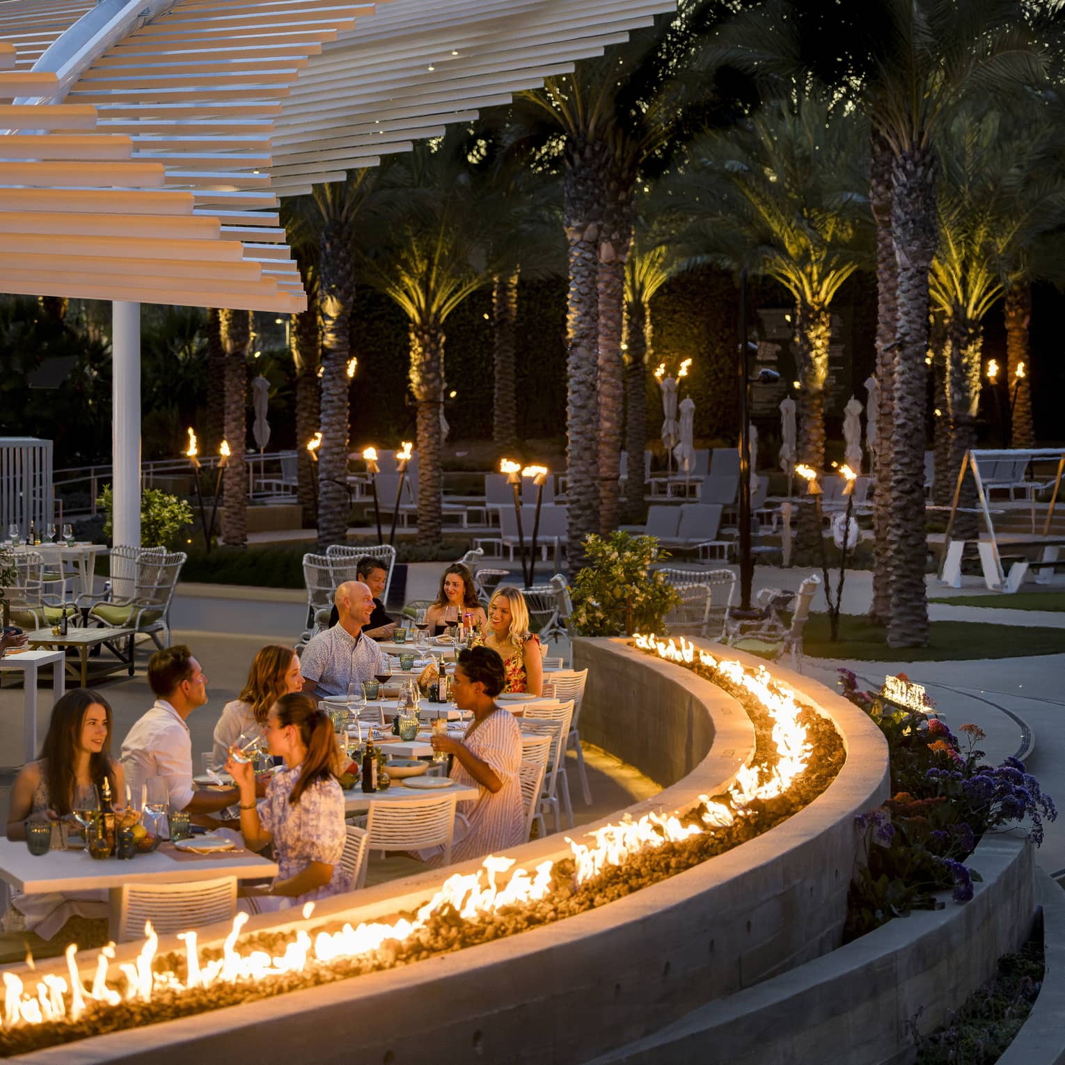 Tables of four enjoying an evening meal on an outdoor patio lined by a curved stone wall topped by a ring of fire