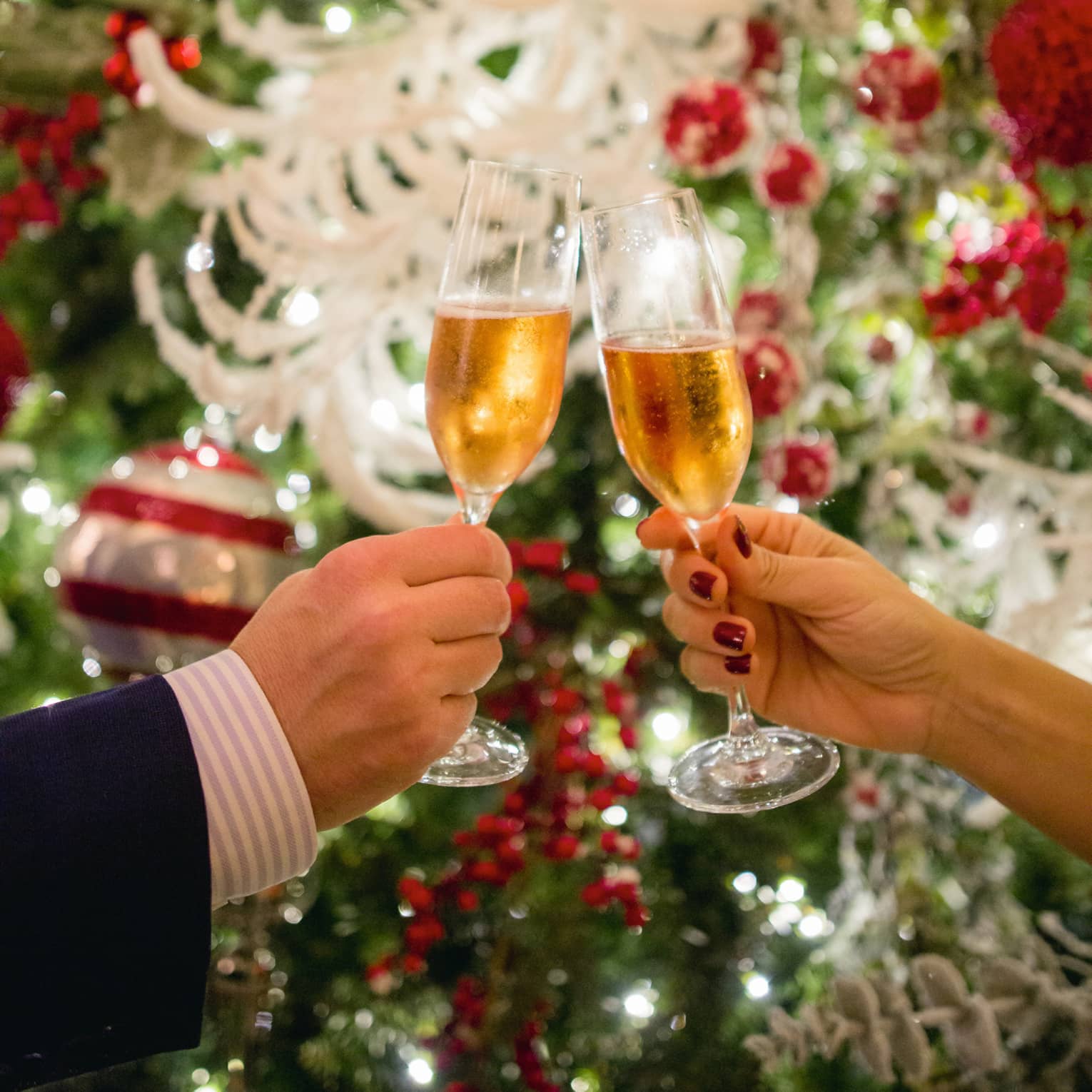 Two hands toasting Champagne glasses with holiday decor in backdrop