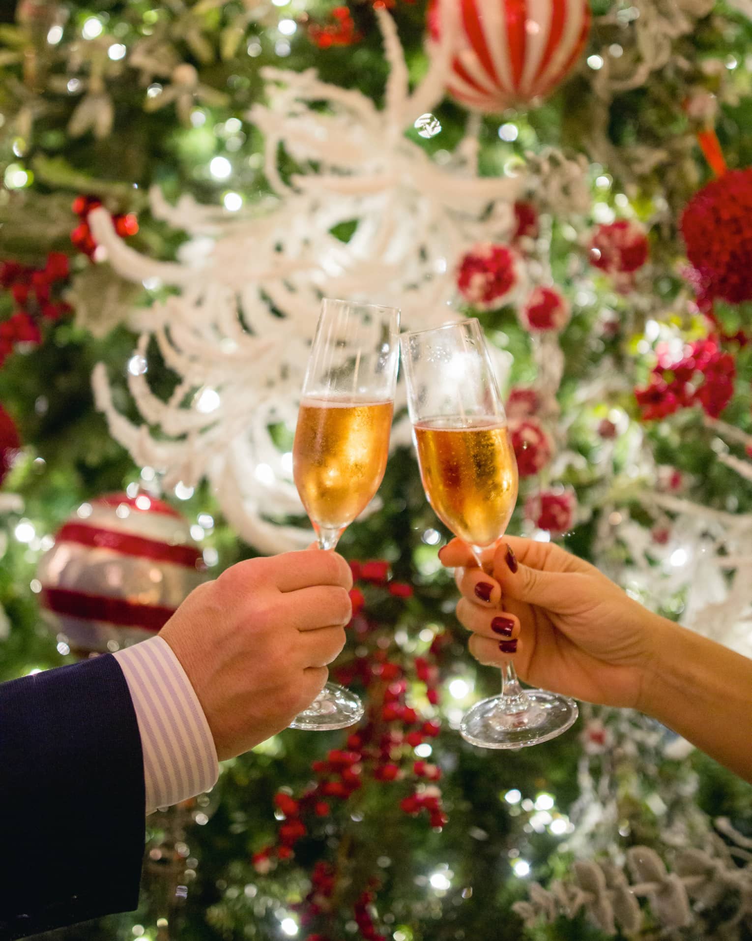 Two hands toasting Champagne glasses with holiday decor in backdrop