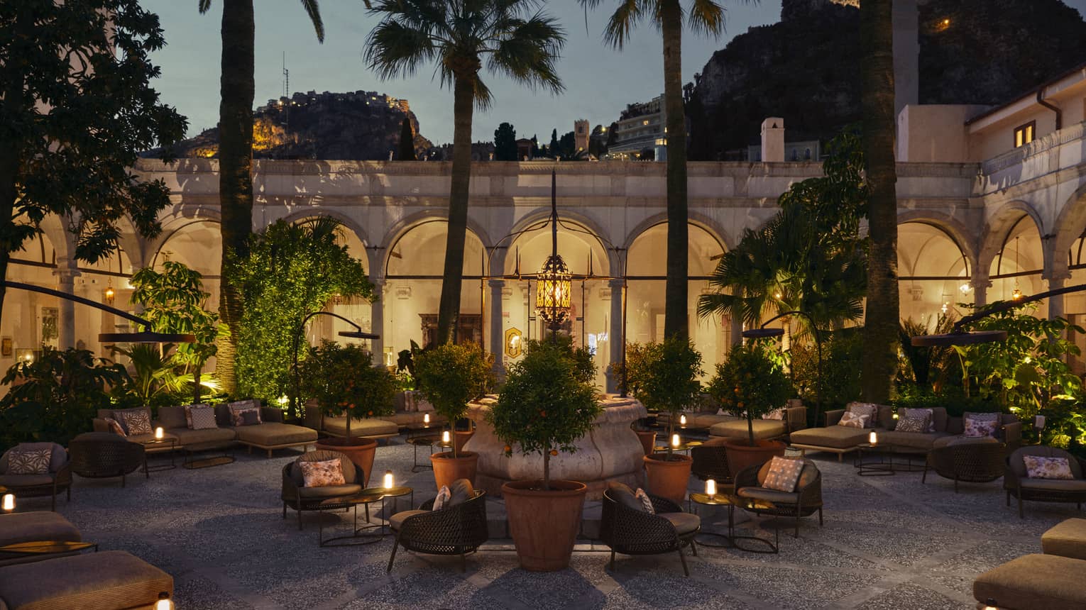 Courtyard-style lounge area with various seating areas and tables topped with candles, all set beneath some trees with an arched colonnade walkway in the background at night