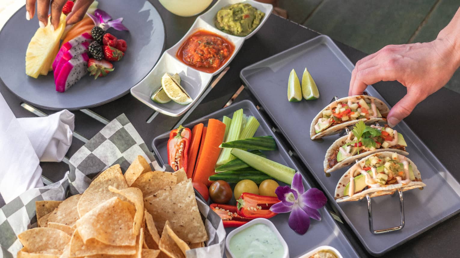 Table with tacos, a fruit plate, vegetables, and chips and salsa.