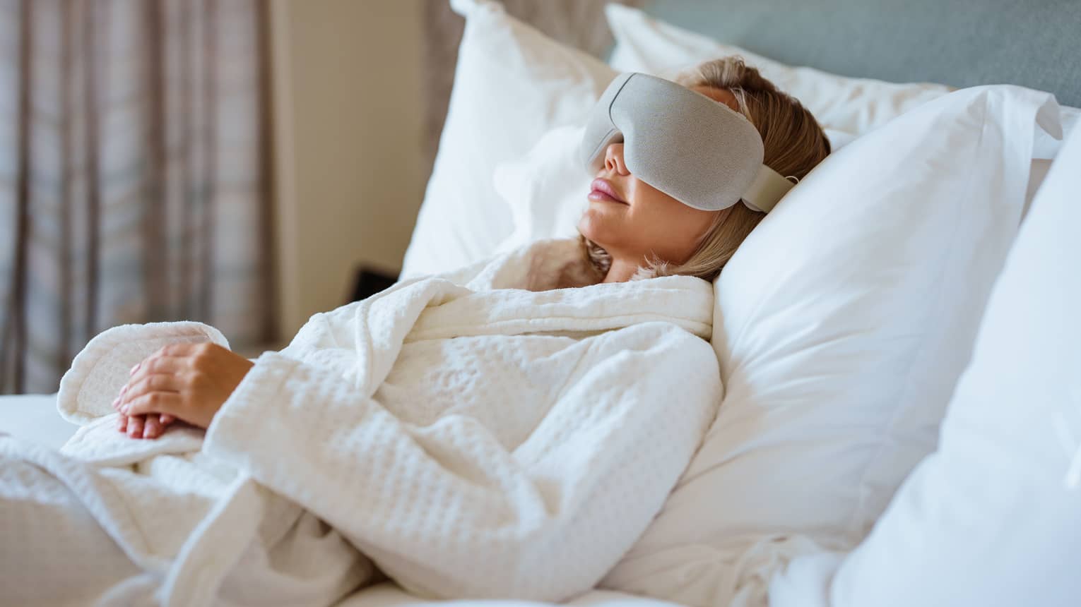 Person wearing white spa robe lays back on a bed with large white pillows wearing a high-tech eye mask