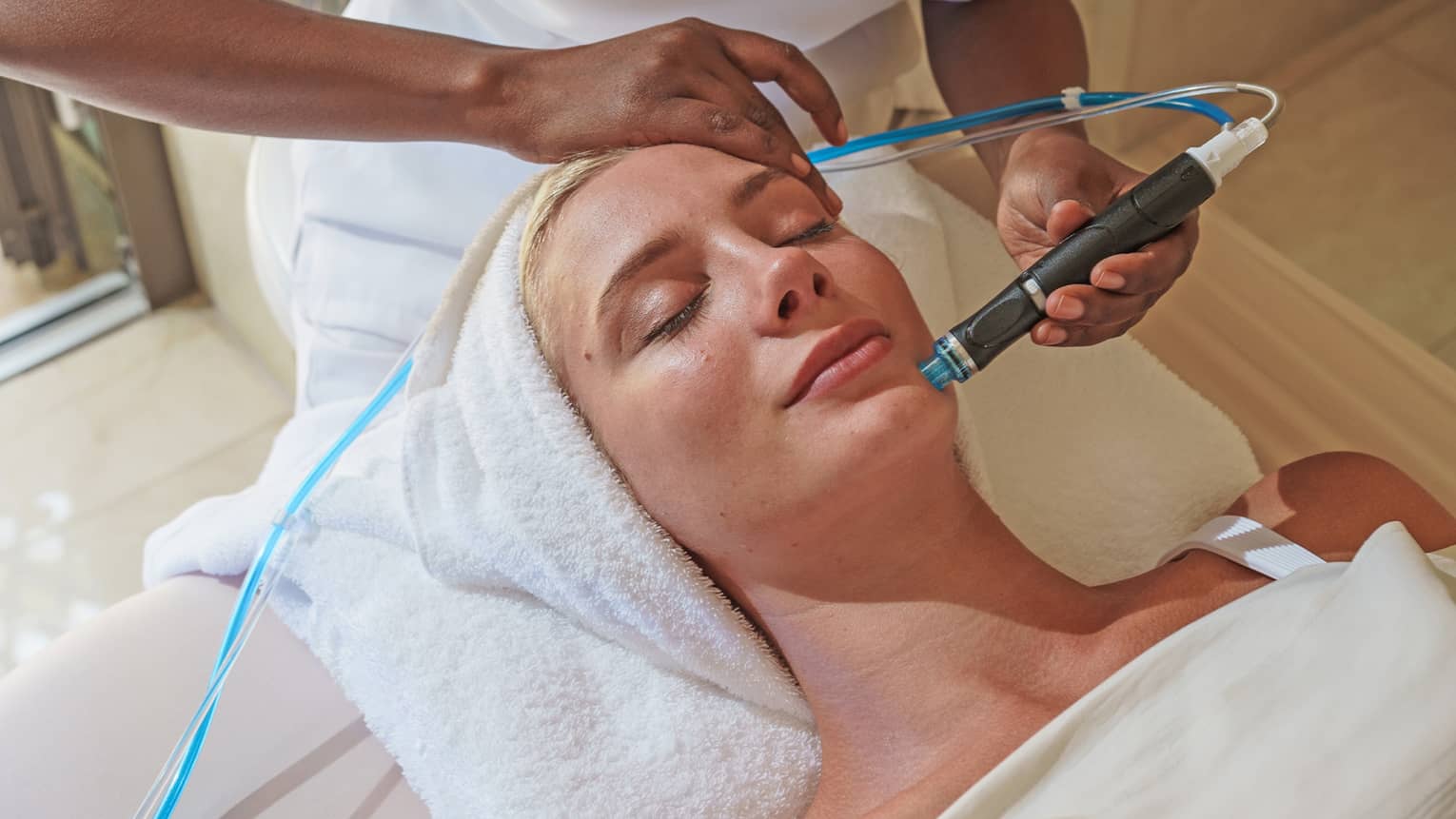 Spa staff holds a blue and black facial tool over woman's face as she lays on massage bed 