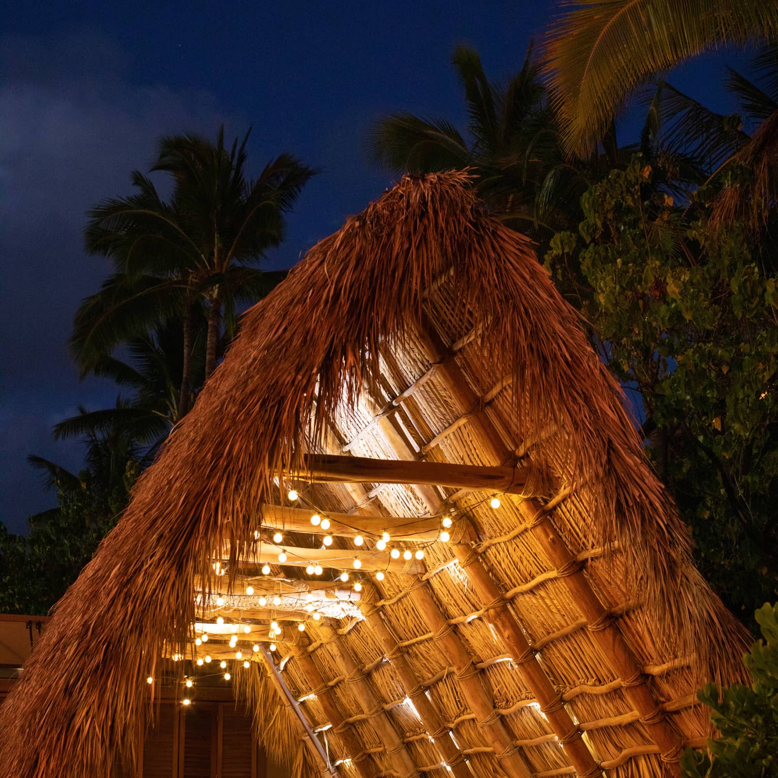 A group dinner setting with candles and twinkling patio lights in a large timber A-frame Hawaiian hale with a thatched roof.