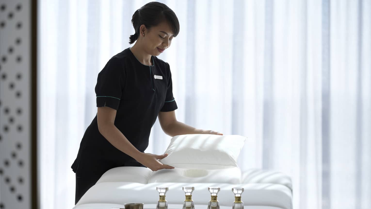 Spa attendant wearing a black uniform fluffs a while pillow on a treatment table covered with all-white linens and a tray filled with four small glass bottles