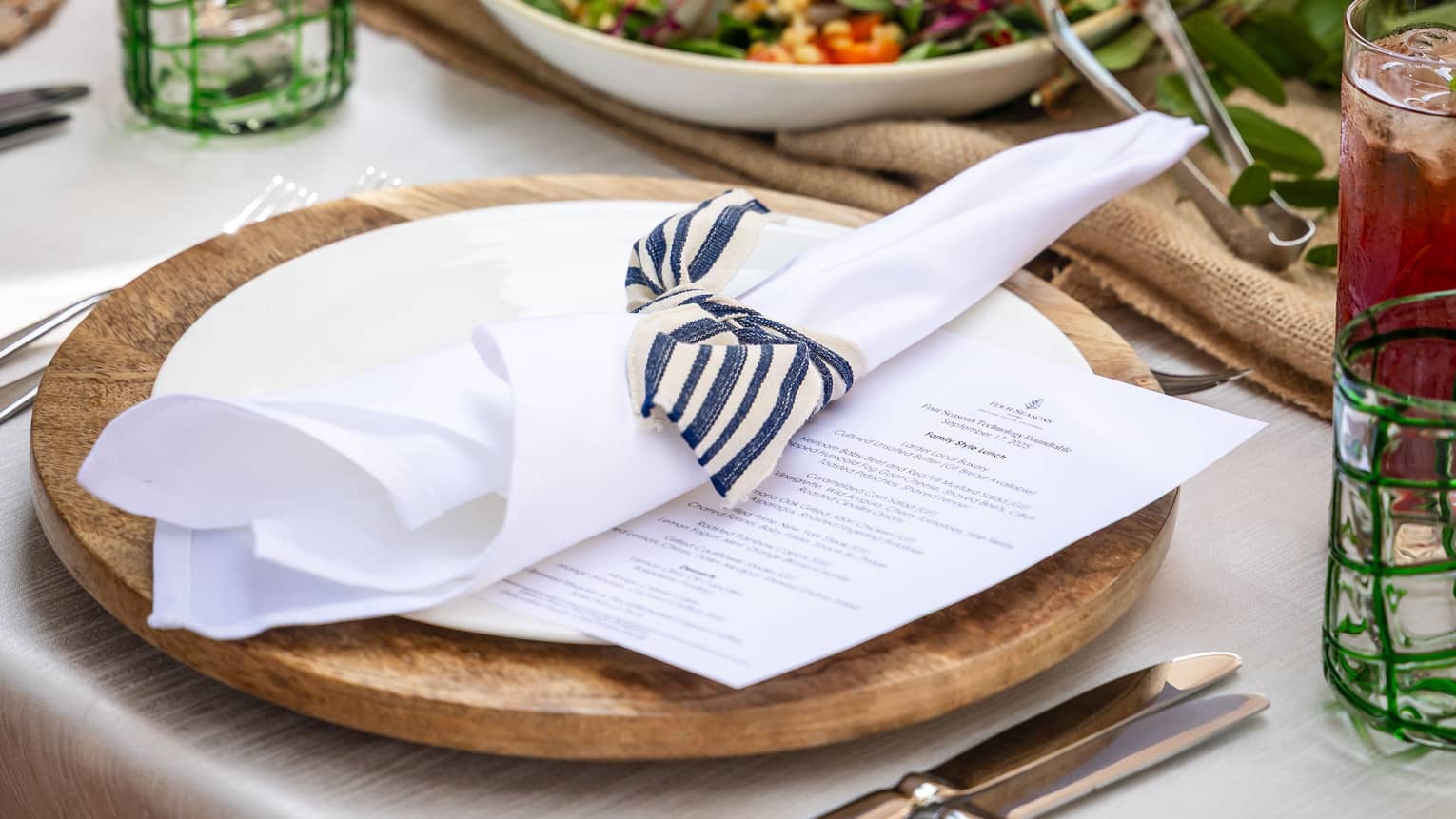 Place setting with a wooden charger, white plate, white napkin tied with a black-and-white striped ribbon sits in front of a large bowl of salad
