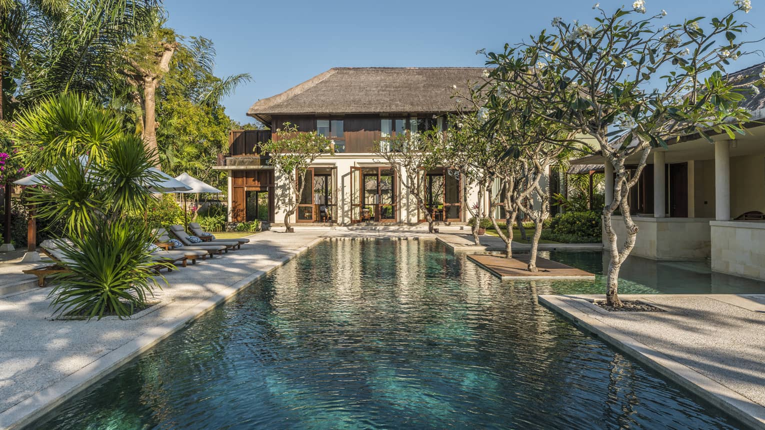 Two-storey villa at end of long outdoor swimming pool lined with trees, tropical plants