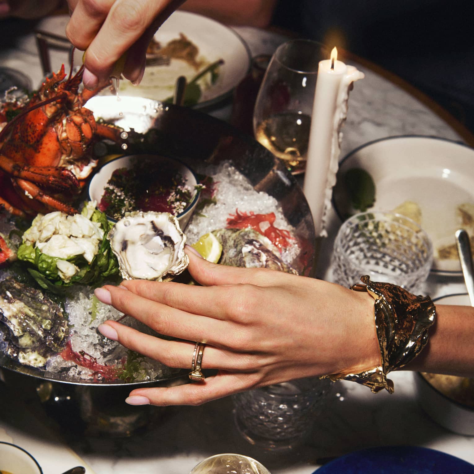 A pair of hand reaches to grab an oyster from a silver seafood platter
