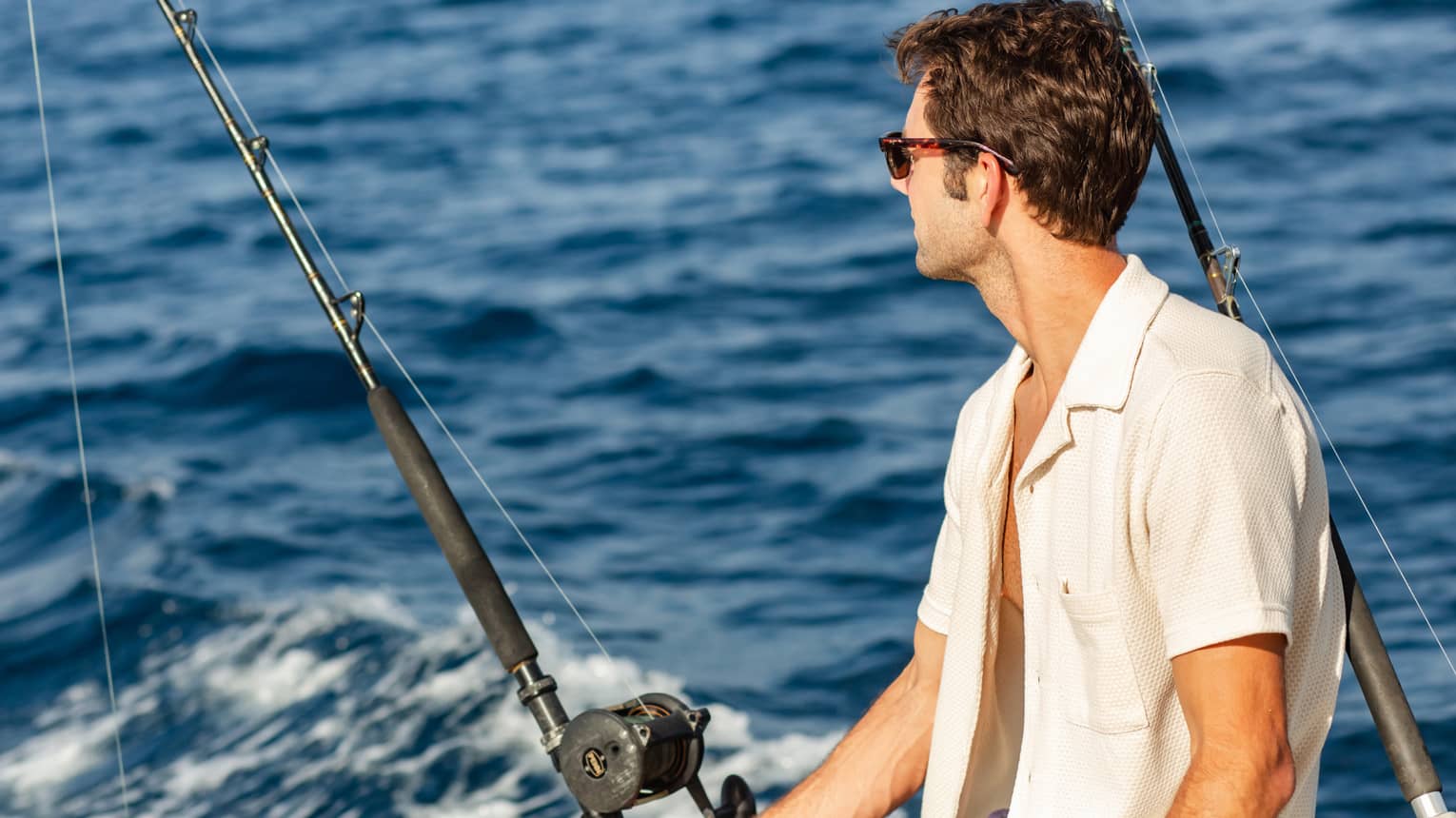 Close-up of a guest wearing sunglasses, sitting beside two offshore fishing rods and looking out towards dark blue waves.