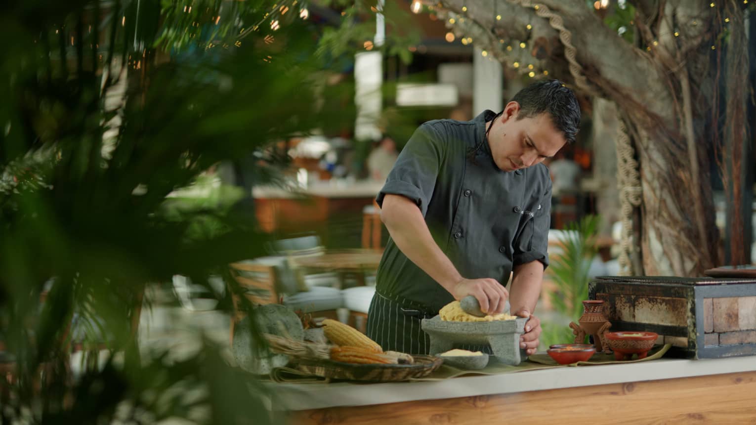 Chef wearing dark grey uniform mashes corn using a stone mortar and pestle