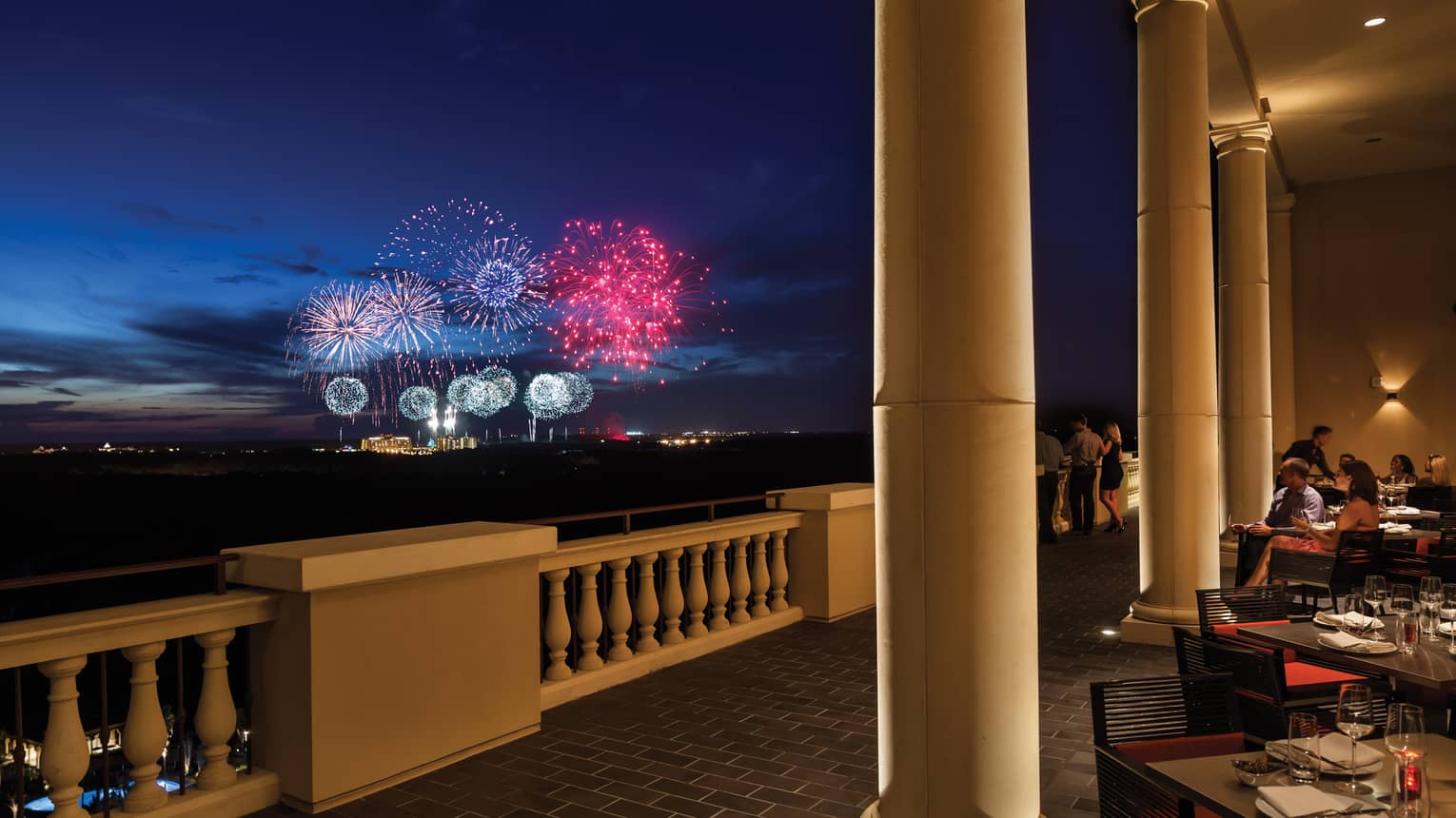 The terrace of a restaurant with tables and people watching fireworks in the distance.