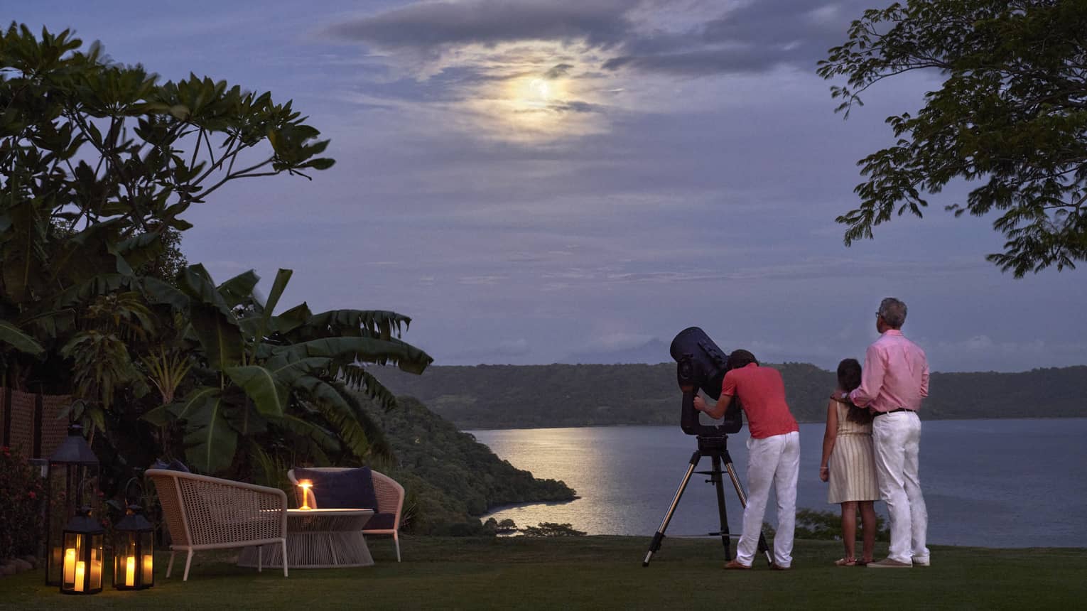 Standing in a treed alcove with a large telescope, a family gazes into the sky, the moon’s glow reflected in the water below.