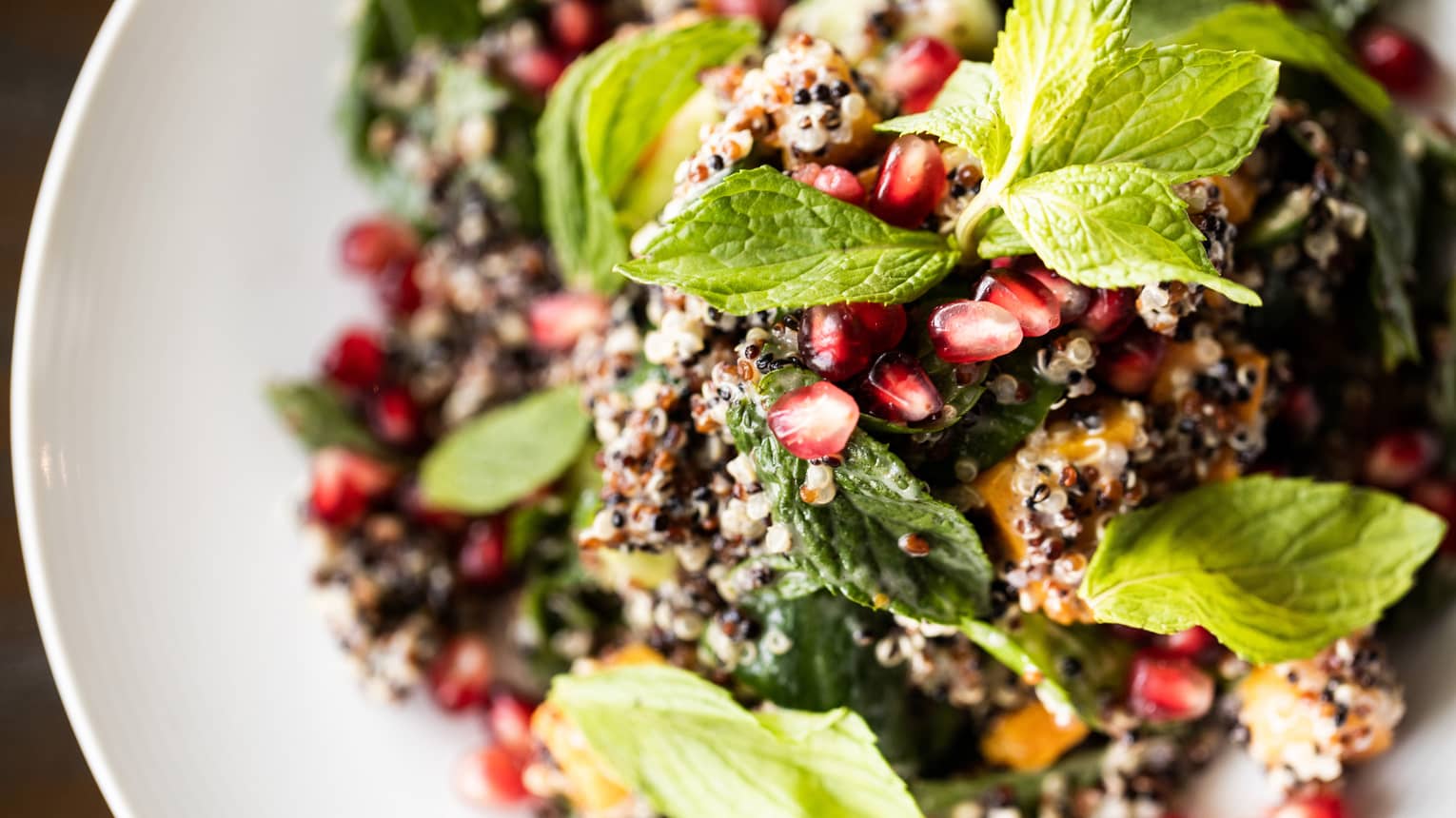 Fresh quinoa salad with green mint leaves, red pomegranate seeds and various vegetables all on a white round plate