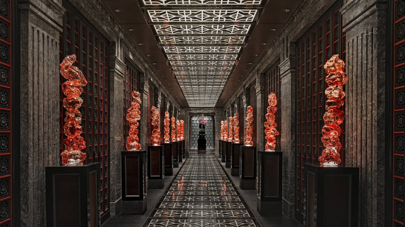 View down a long black-tiled hall lined with orange glass sculptures on pedestals, the geometric ceiling mirroring the floor.