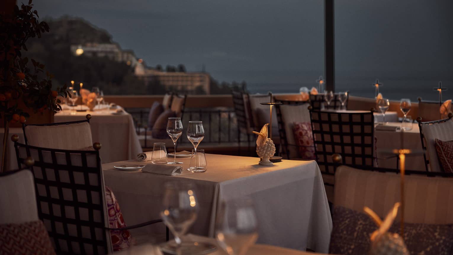 Various tables each topped with white tablecloths, wine glasses and small illuminated lights fill a restaurant terrace overlooking the sea at night