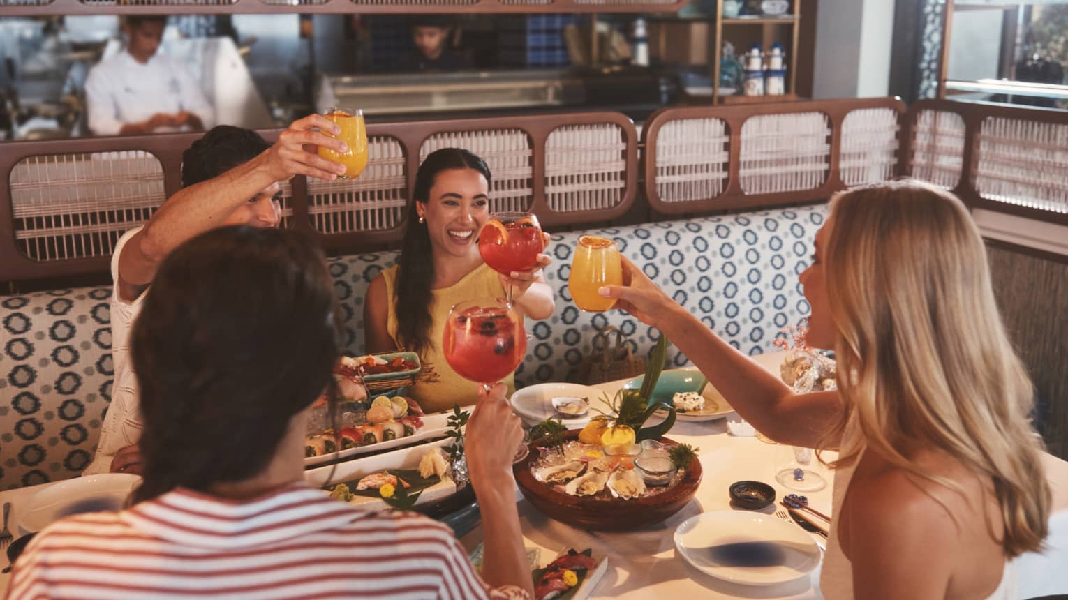 Four people sit in an blue-and-white upholstered restaurant booth, each raising a glass and smiling over a table filled with seafood platters