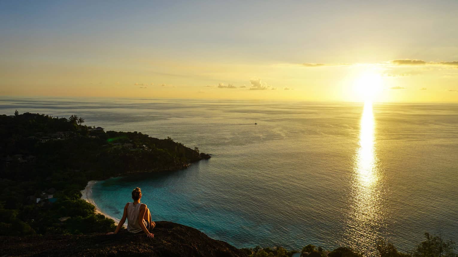 Woman sits on cliff looking out at sunset over ocean