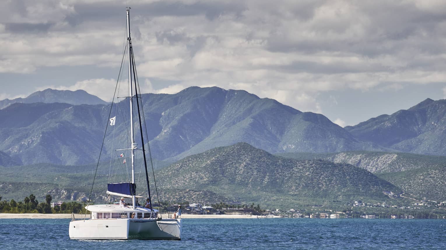 A catamaran on calm, blue water under a partly-cloudy sky with low green hills in the background and mountain ranges beyond.