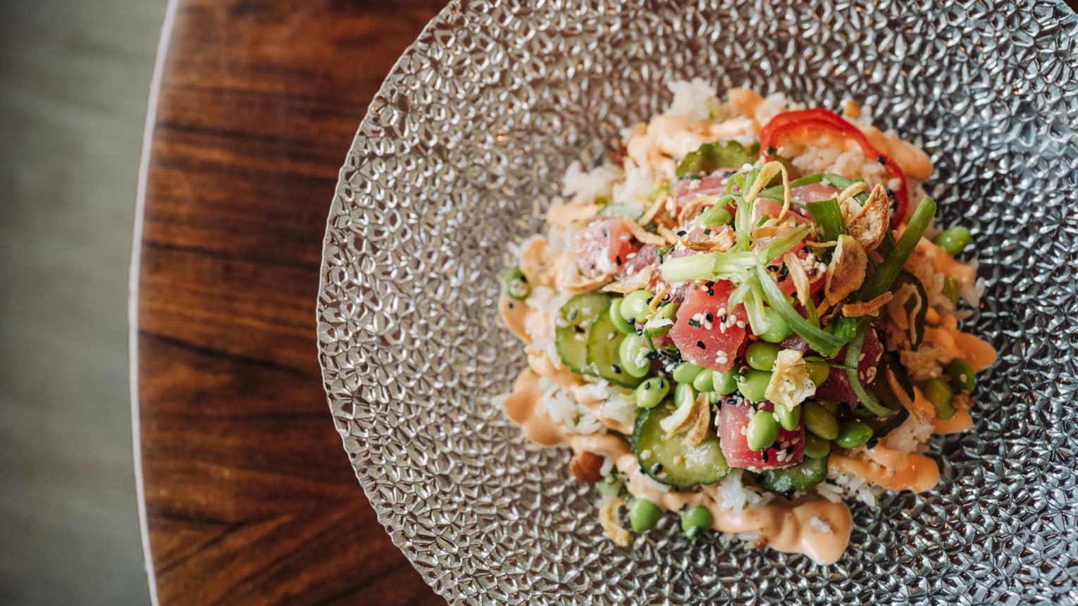 Tuna poke with pickled ginger, candied cashew rice, cucumber, crispy shallots, spicy aioli and sesame seeds served on a clear glass plate