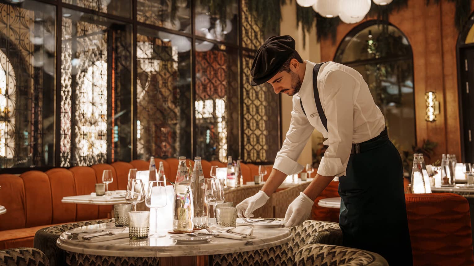 Waiter wearing white button-down shirt, black pants, black suspenders, black beret and white gloves arranges a plate on a table in an Art Deco?style restaurant