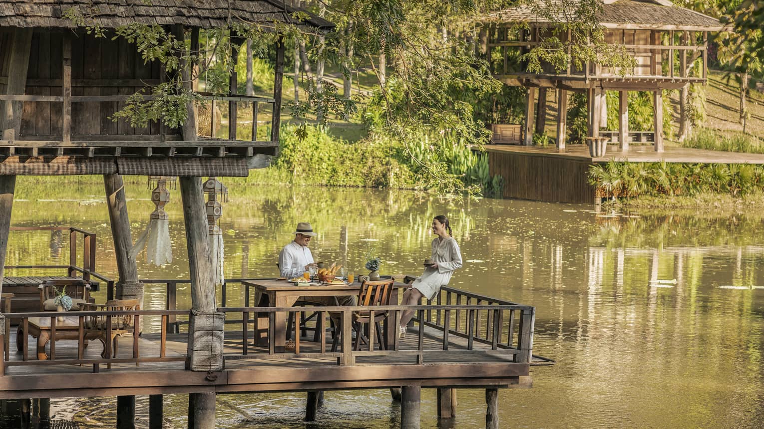 Two guests at a rustic table, on a platform in one of two wood huts on stilts perched atop a calm river bordered by greenery.