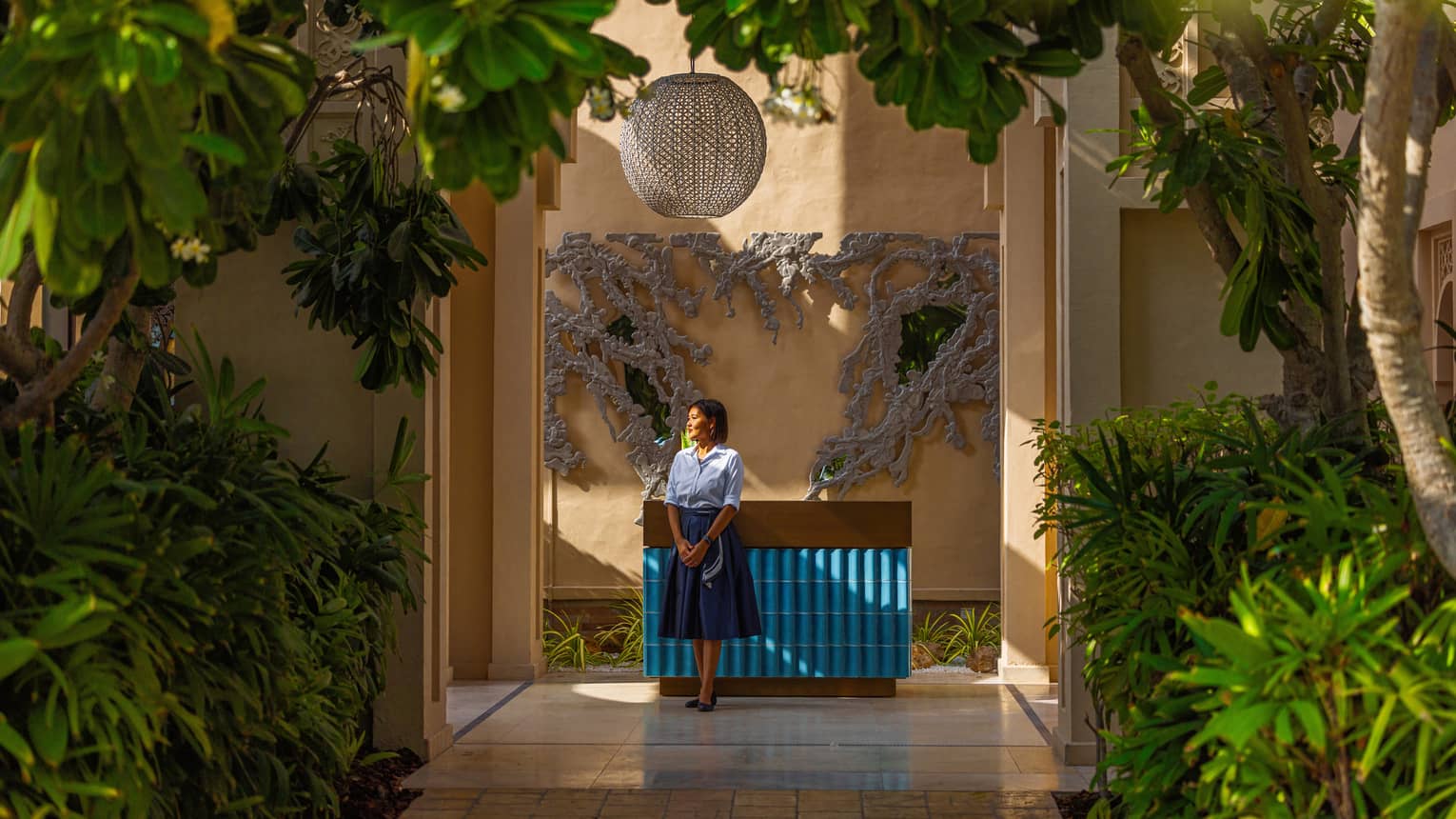 Woman staff member wearing light-blue button down shirt and dark blue knee-length skirt stande next to a blue-tiled counter set surrounded by bright green plants and trees