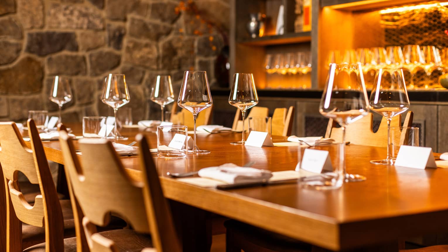 Table set for six in a dining room with a stone walls and a shelf filled with wine glasses