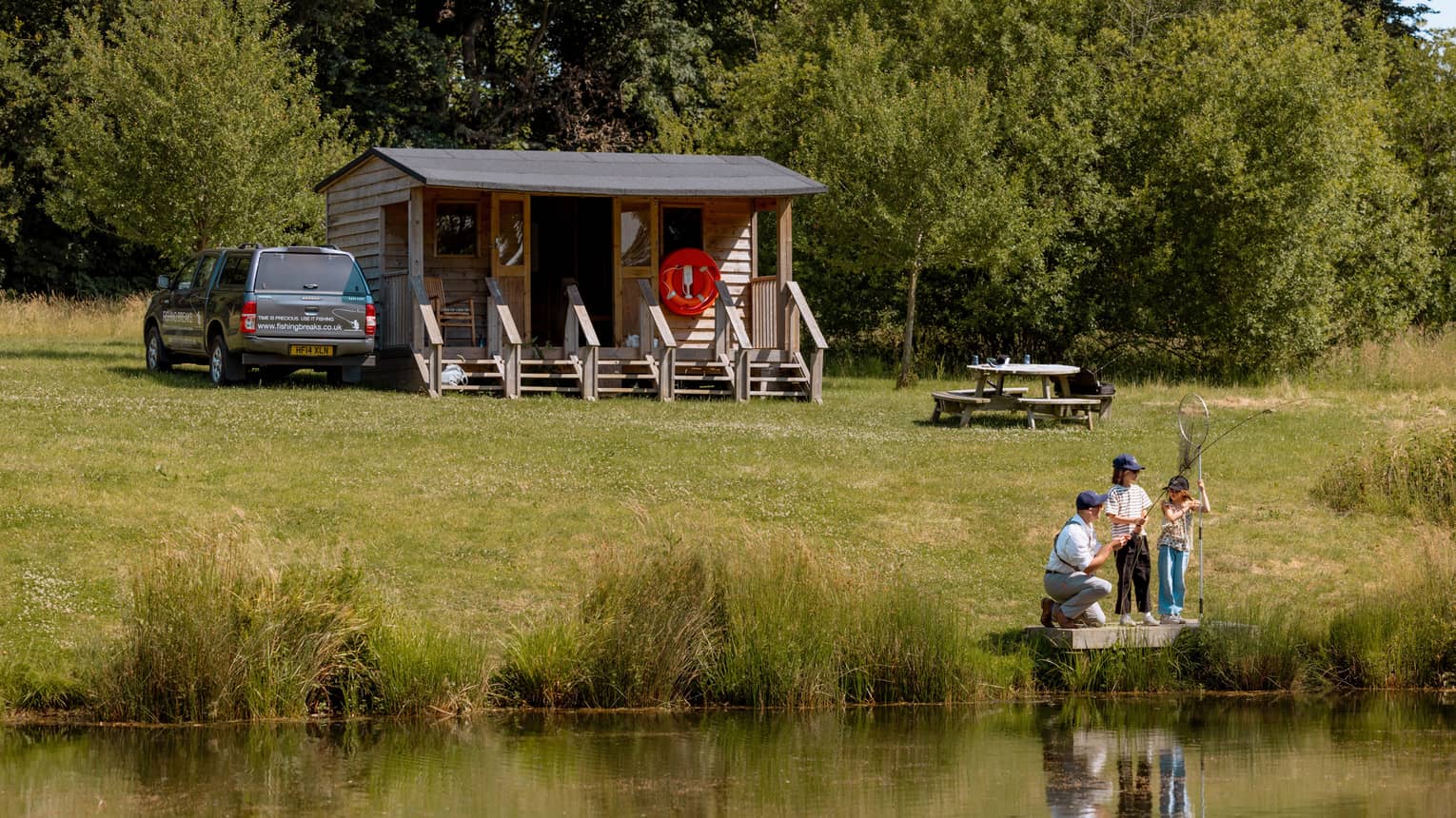 Three people fishing on small platform by lake with cabin-like building and picnic table in backdrop