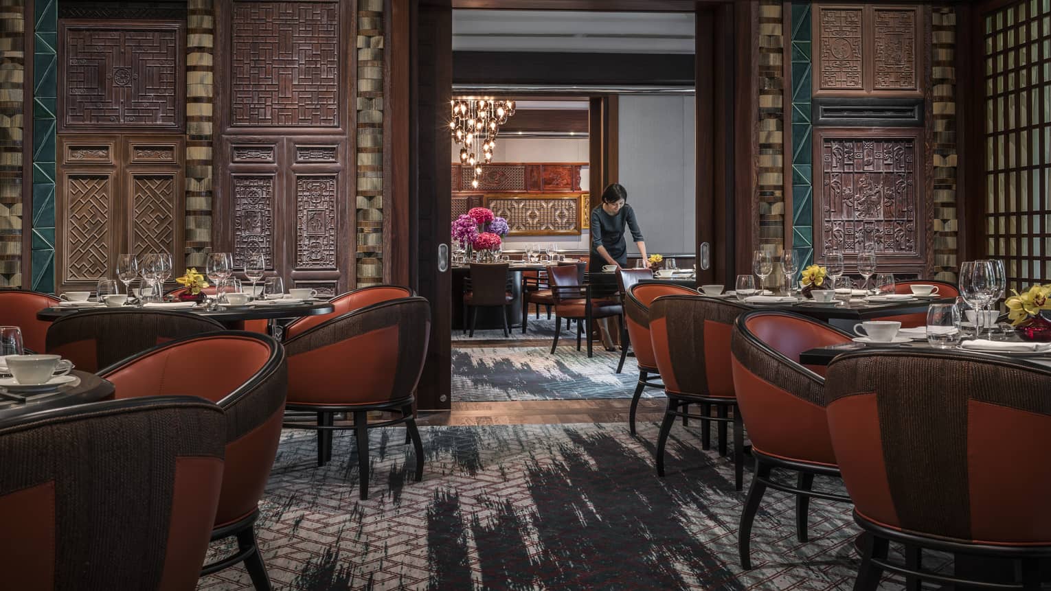 Woman sets table at Jiang-Nan Chun, Michelin-starred Cantonese restaurant, with red leather bucket chairs around dining tables, carved wood walls.