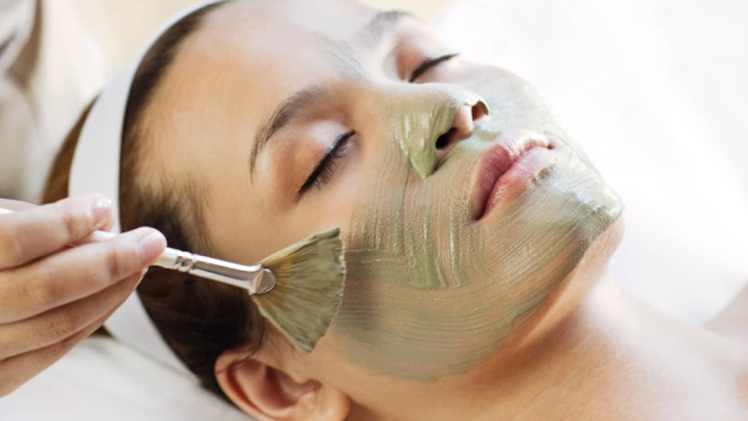 Spa staff holding brush paints green mask on woman's cheeks, chin, nose 