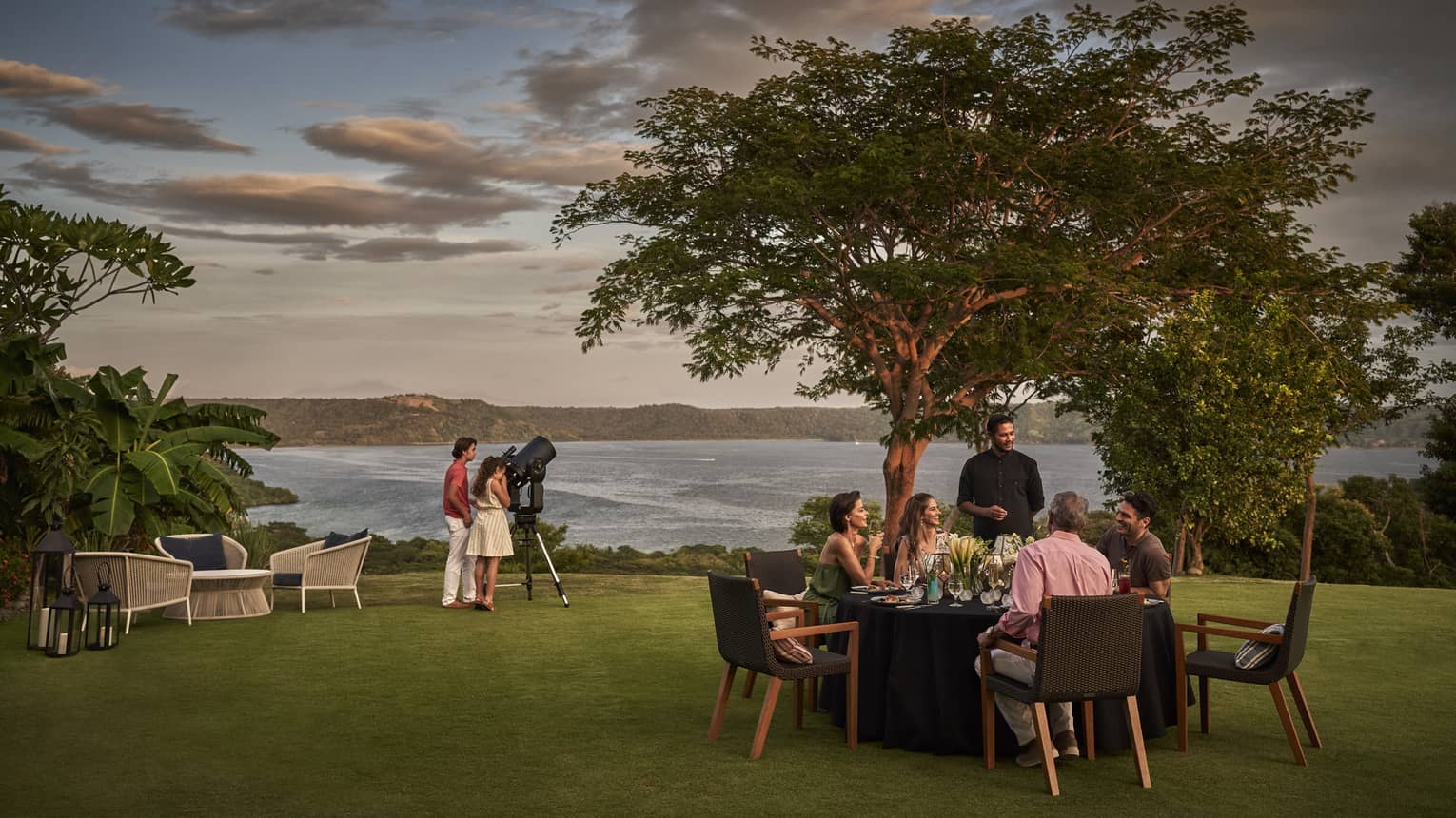 Group of people sit at a private dining table set on a lawn overlooking the ocean while two others look through a telescope